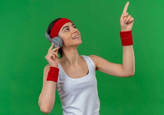 Young fitness woman in sportswear with headband and headphones smiling cheerfully pointing up with index finger standing over green wall