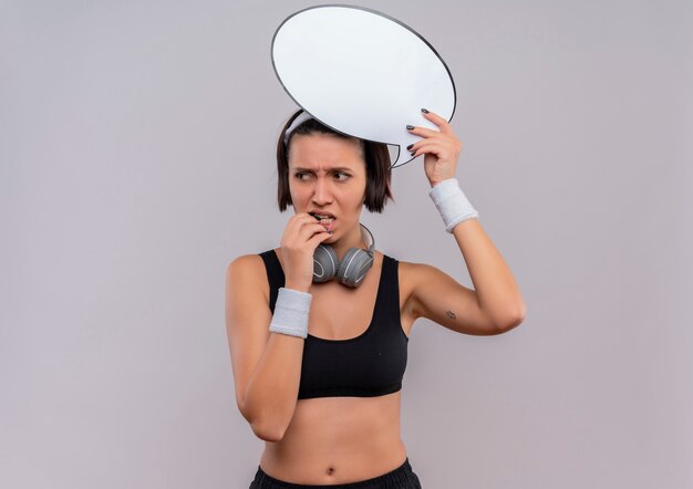 Молодая фитнес-женщина в спортивной одежде с повязкой на голову, держащая пустой речевой пузырь, озадаченно смотрит в сторону, стоя над белой стеной