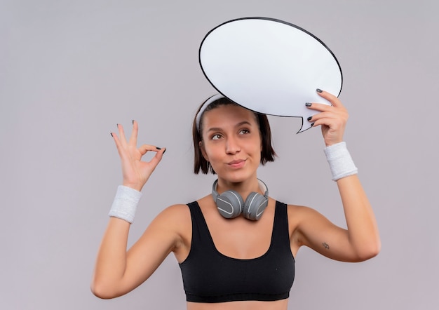 Young fitness woman in sportswear with headband holding blank speech bubble sign making meditation gesture with fingers feeling positive emotions smiling standing over white wall