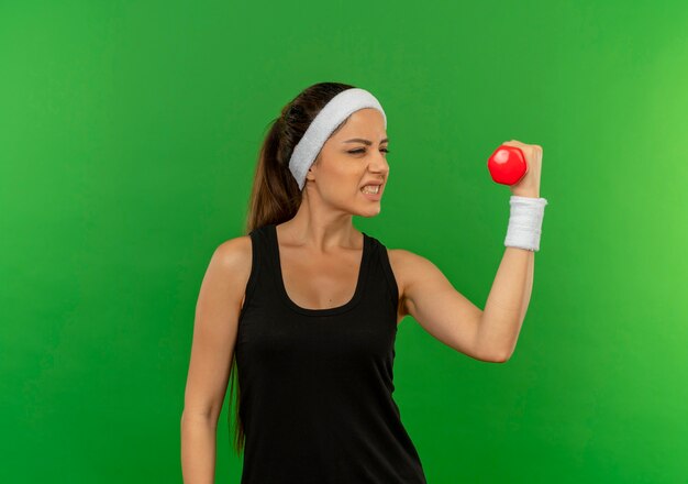 Молодая женщина фитнеса в спортивной одежде с повязкой на голову, держащей гантели, делает упражнения, выглядя напряженными, стоя над зеленой стеной