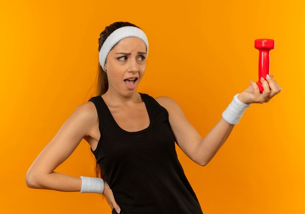 Young fitness woman in sportswear with headband holding dumbbell looking at it confused standing over orange wall