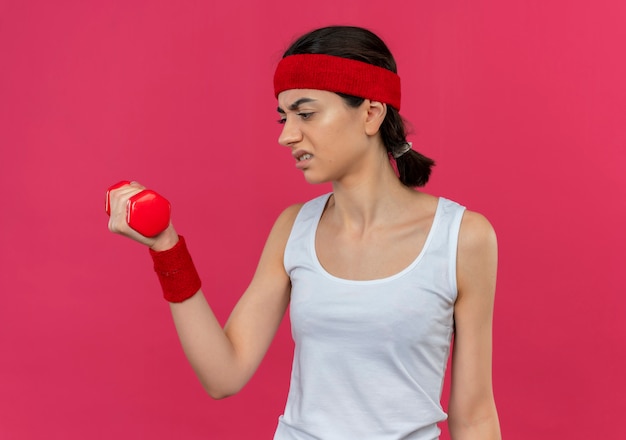 Young fitness woman in sportswear with headband holding dumbbell in raised hand looking at it with annoyed expression on face standing over pink wall