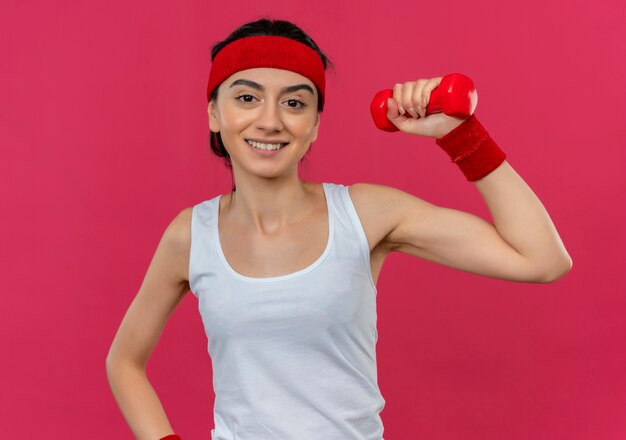 Young fitness woman in sportswear with headband holding dumbbell in raised hand smiling confident standing over pink wall