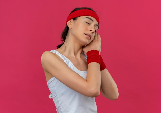 Молодая фитнес-женщина в спортивной одежде с повязкой на голову, держащая ладони вместе с закрытыми глазами, положив голову на ладони, хочет спать, стоя у розовой стены