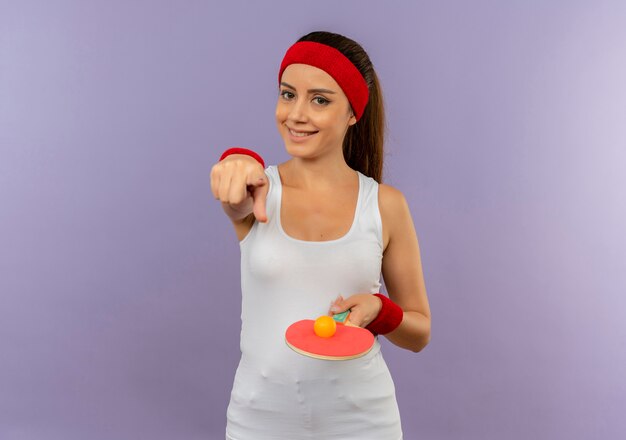 Young fitness woman in sportswear with headband holding racket and ball for table tennis pointing with index finger to camera with smile on face standing over grey wall