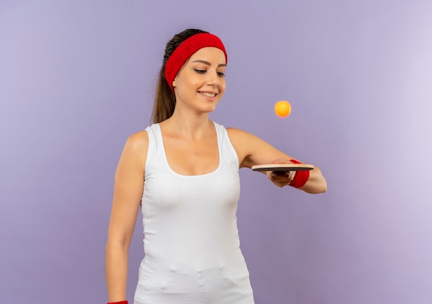 Giovane donna fitness in abiti sportivi con fascia tenendo la racchetta e la palla per ping-pong sorridente allegramente in piedi sopra il muro grigio