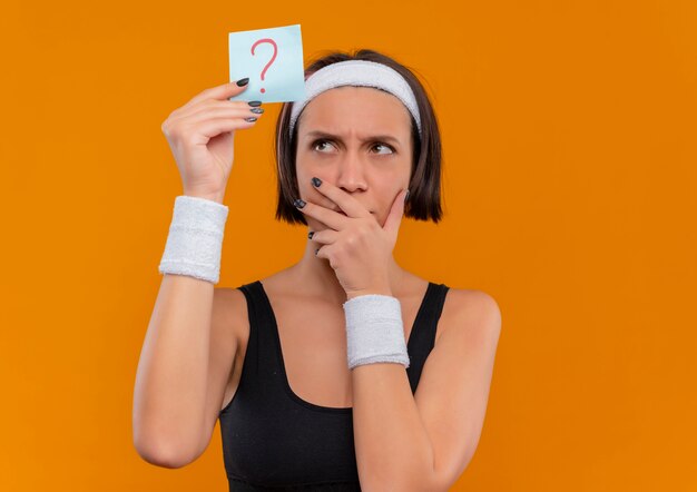 Young fitness woman in sportswear with headband holding reminder paper with question mark looking at it with pensive expression thinking standing over orange wall