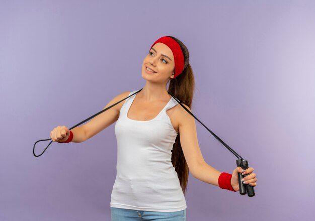 Young fitness woman in sportswear with headband holding skipping rope happy and positive smiling cheerfully standing over grey wall