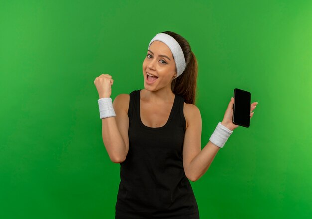 Young fitness woman in sportswear with headband holding smartphone clenching fist smiling cheerfully standing over green wall