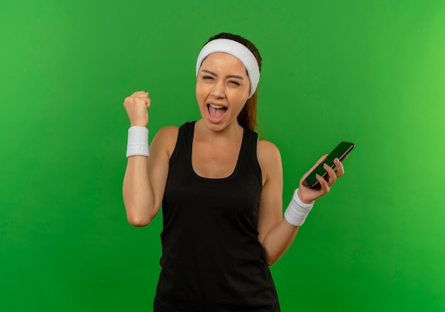 Young fitness woman in sportswear with headband holding smartphone raising fist happy and excited standing over green wall