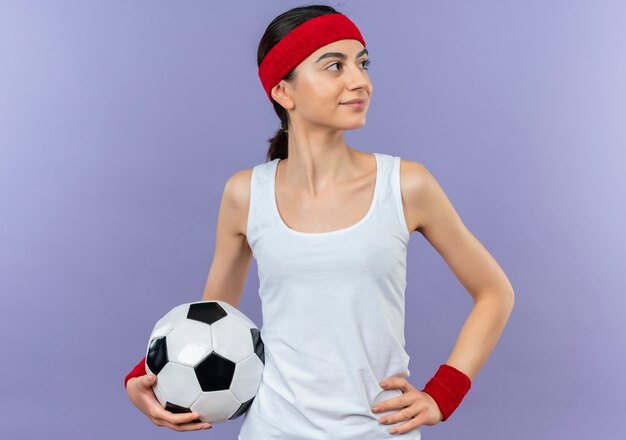Молодая женщина фитнеса в спортивной одежде с повязкой на голову держит футбольный мяч, глядя в сторону с уверенной улыбкой на лице, стоящем над фиолетовой стеной