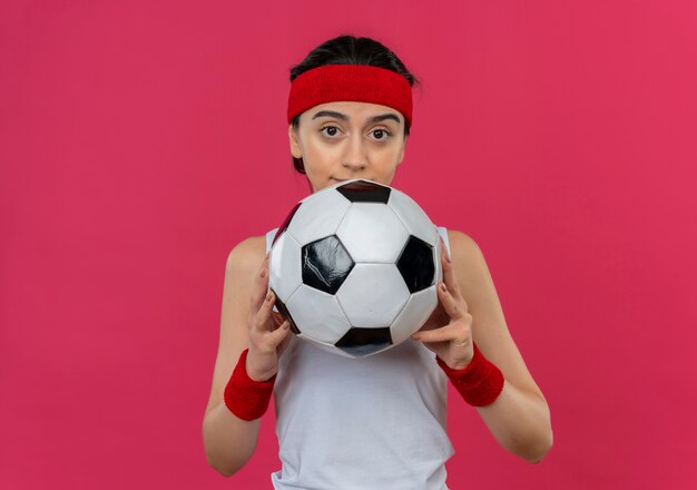 Young fitness woman in sportswear with headband holding soccer ball looking confused at camera standing over pink wall