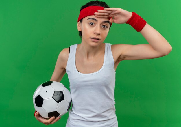 緑の壁の上に立っている頭の上に手で遠くを見ているサッカーボールを保持しているヘッドバンドを持つスポーツウェアの若いフィットネス女性