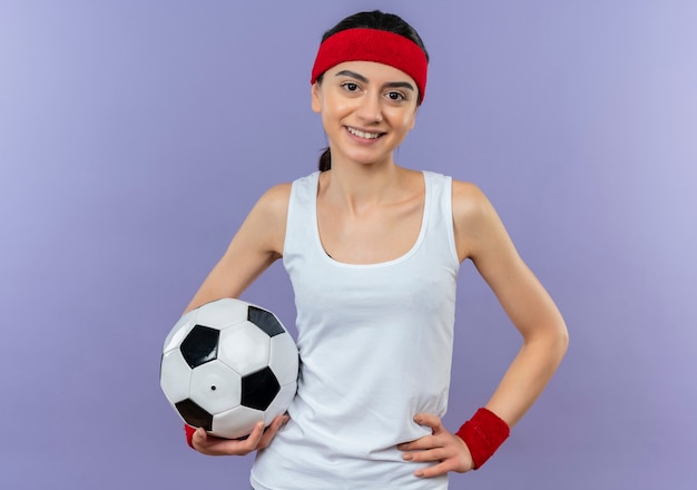 Молодая фитнес-женщина в спортивной одежде с повязкой на голову, держащая футбольный мяч, уверенно улыбается, счастлива и позитивно стоит над фиолетовой стеной