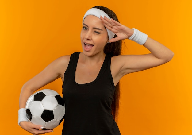 Молодая женщина фитнеса в спортивной одежде с повязкой на голову держит футбольный мяч, подмигивая и улыбаясь, отдавая честь стоя над оранжевой стеной