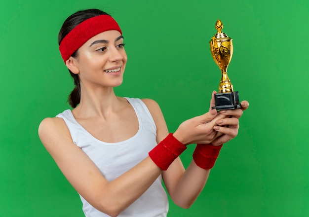 Giovane donna fitness in abiti sportivi con fascia tenendo il trofeo guardandolo sorridente fiducioso in piedi sopra la parete verde