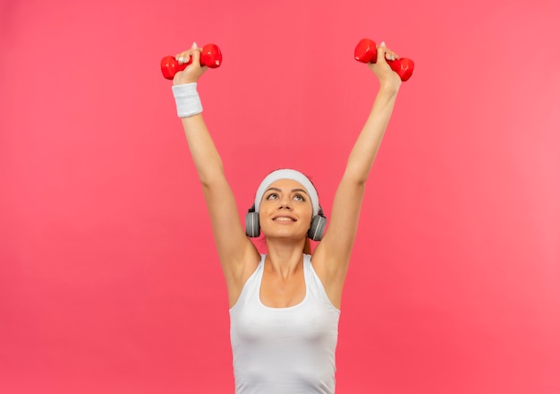 Young fitness woman in sportswear with headband holding two dumbbells doing exercises looking confident standing over pink wall