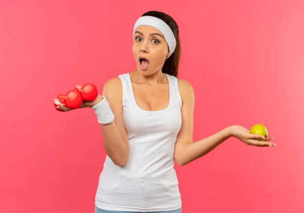 Young fitness woman in sportswear with headband holding two dumbbells and green apple looking surprised and amazed standing over pink wall