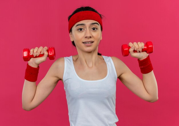 Молодая фитнес-женщина в спортивной одежде с повязкой на голову, держащая две гантели в поднятых руках, смущенная, стоя над розовой стеной