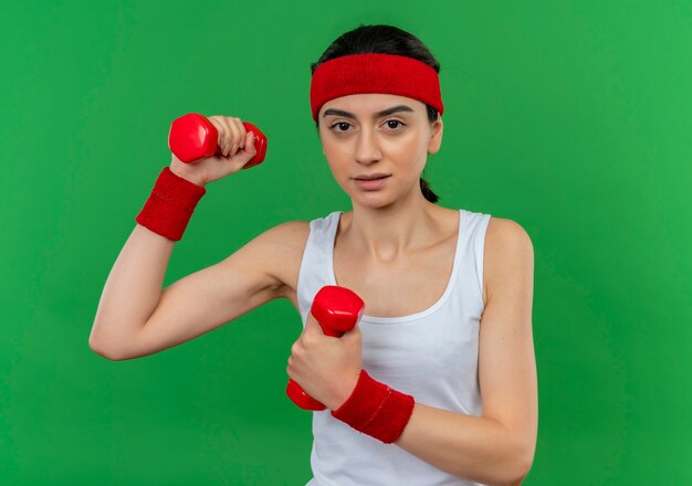 Young fitness woman in sportswear with headband holding two dumbbells raising hands doing exercises confident with serious face standing over green wall