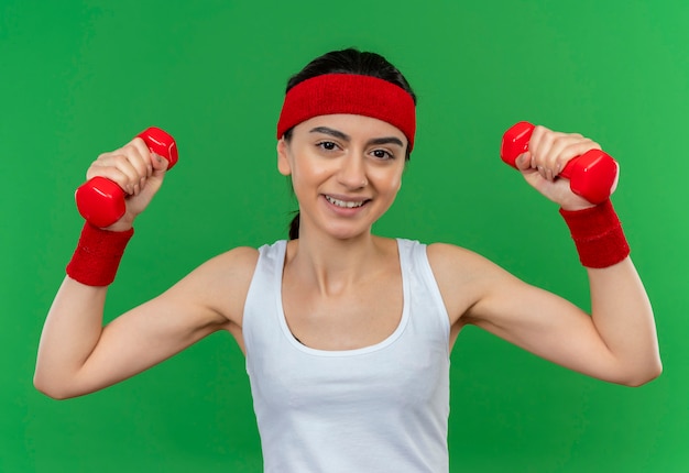 Young fitness woman in sportswear with headband holding two dumbbells raising hands doing exercises smiling confident standing over green wall