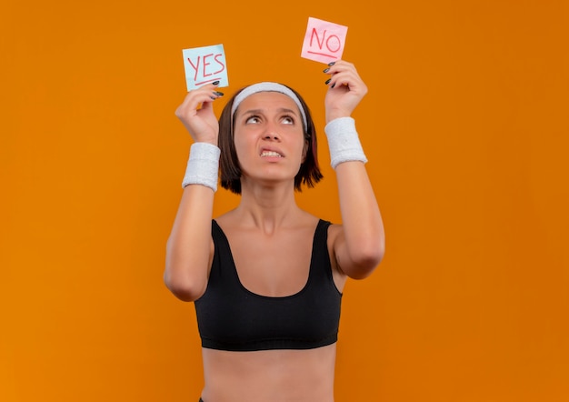 Free photo young fitness woman in sportswear with headband holding two reminder papers with word yes and no in raised hands looking at them confused standing over orange wall