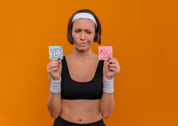 Foto gratuita giovane donna fitness in abbigliamento sportivo con fascia tenendo due fogli di promemoria con la parola sì e no in mani alzate guardandoli con espressione scettica in piedi sopra la parete arancione