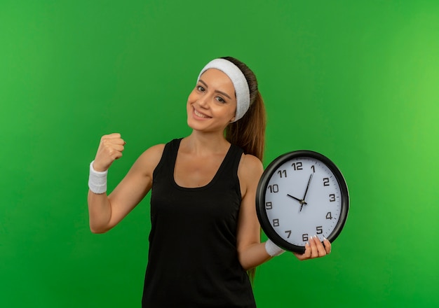 Giovane donna fitness in abiti sportivi con fascia tenendo l'orologio da parete stringendo il pugno felice e positivo sorridente in piedi sopra la parete verde