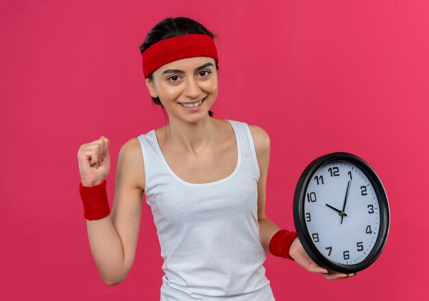 Young fitness woman in sportswear with headband holding wall clock clenching fist smiling cheerfully happy and positive standing over pink wall