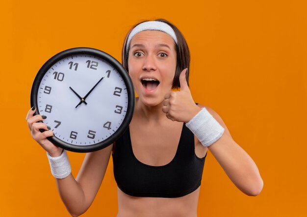 Молодая фитнес-женщина в спортивной одежде с повязкой на голову, держащая настенные часы, счастливая и взволнованная, показывает палец вверх, стоя над оранжевой стеной
