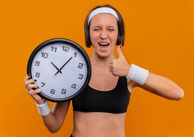 Young fitness woman in sportswear with headband holding wall clock happy and excited showing thumbs up standing over orange wall