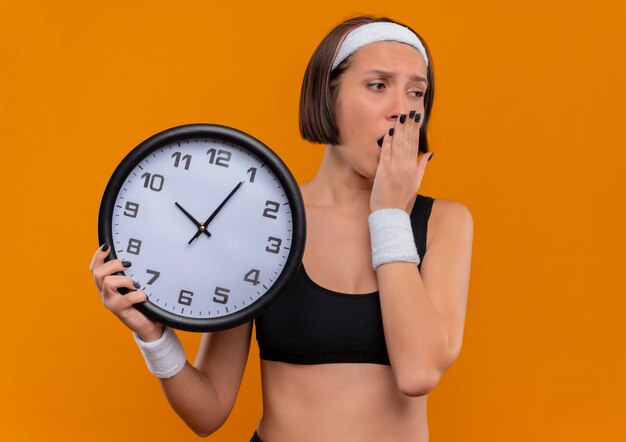 Молодая фитнес-женщина в спортивной одежде с повязкой на голову, держащая настенные часы, выглядит усталой и скучающей, зевая, стоя над оранжевой стеной