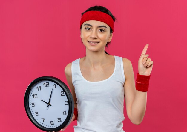 Молодая фитнес-женщина в спортивной одежде с повязкой на голову, держащая настенные часы, показывающая, что указательный палец выглядит уверенно, стоит над розовой стеной
