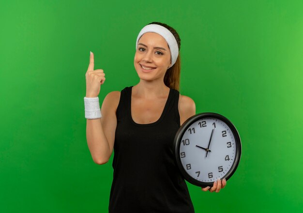 Young fitness woman in sportswear with headband holding wall clock showing index finger smiling confident standing over green wall