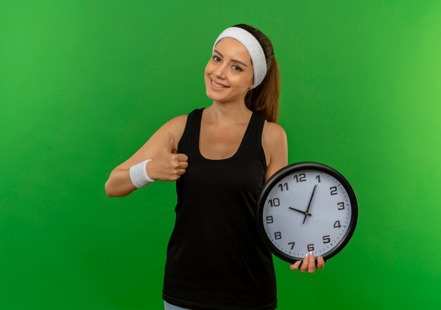 Young fitness woman in sportswear with headband holding wall clock showing thumbs up smiling standing over green wall