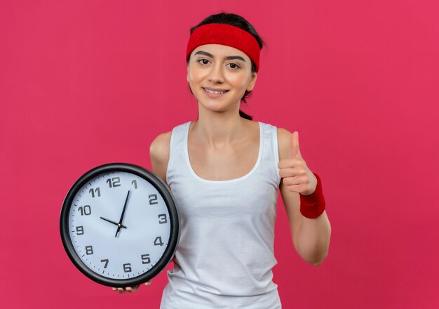 Young fitness woman in sportswear with headband holding wall clock smiling showing thumbs up standing over pink wall