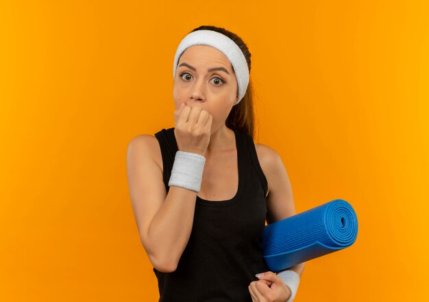Молодая фитнес-женщина в спортивной одежде с повязкой на голову, держащая коврик для йоги, потрясена, прикрывая рот рукой, стоящей над оранжевой стеной