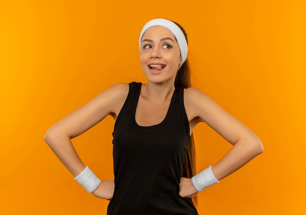 Young fitness woman in sportswear with headband looking aside happy and positive smiling standing over orange wall