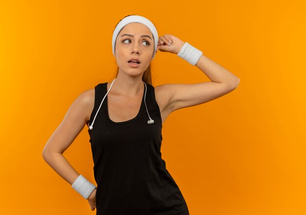 Young fitness woman in sportswear with headband looking aside puzzled standing over orange wall