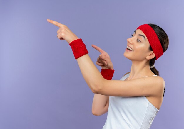 Young fitness woman in sportswear with headband looking aside surprised and amazed pointing with finger to the side standing over purple wall