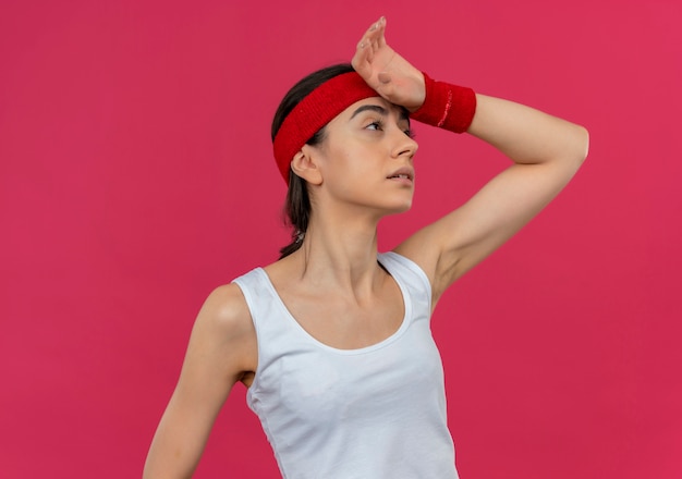 Young fitness woman in sportswear with headband looking aside with hand on head tired after workout standing over pink wall