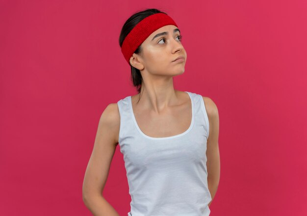 Young fitness woman in sportswear with headband, looking aside with skeptic expression standing over pink wall