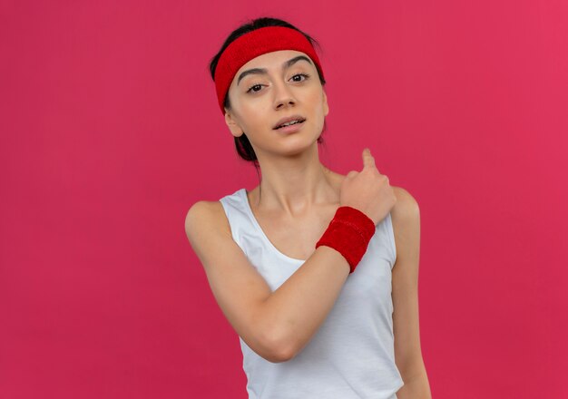 Young fitness woman in sportswear with headband looking confident pointing back standing over pink wall
