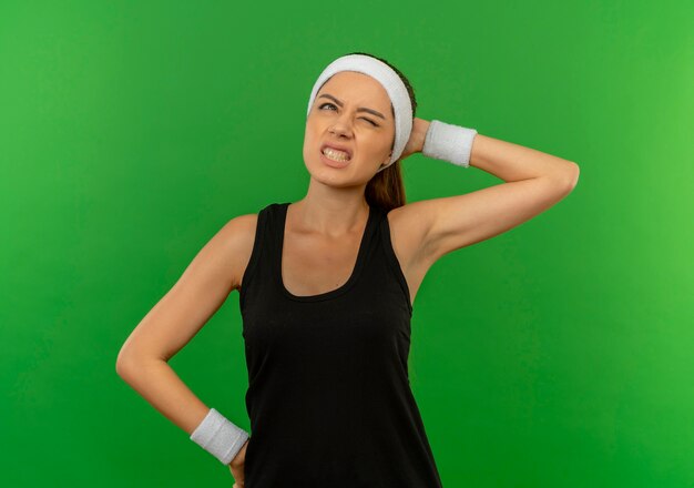 Young fitness woman in sportswear with headband looking confused touching her head for mistake standing over green wall