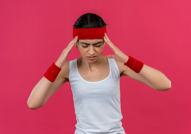 Young fitness woman in sportswear with headband looking tired touching her temples suffering from headache standing over pink wall