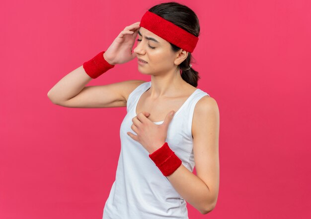Молодая женщина фитнеса в спортивной одежде с повязкой на голову, выглядящая нездоровой, трогательно голова с головной болью, стоя над розовой стеной