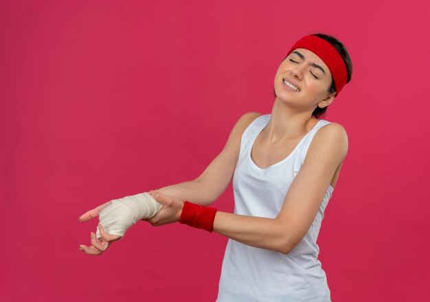 Young fitness woman in sportswear with headband looking unwell touching her bandaged wrist having pain standing over pink wall
