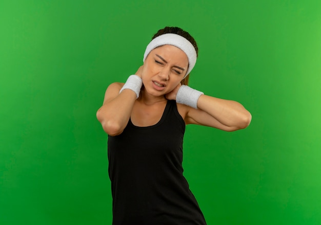 Free photo young fitness woman in sportswear with headband looking unwell touching her neck having pain standing over green wall