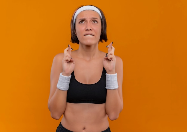 Young fitness woman in sportswear with headband making desirable wish crossing fingers with hope expression standing over orange wall