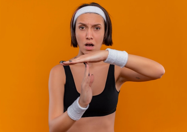 Foto gratuita giovane donna fitness in abiti sportivi con fascia che fa il gesto di time out che sembra confuso in piedi sopra la parete arancione